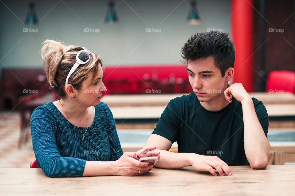 Woman and young man talking together and using mobile phones sitting by a table in cafe