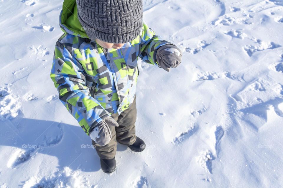 A child with a serious expression on his face in winter clothes jackets, pants, hat and boots in winter on the white snow on the street and in the park in nature plays winter fun.