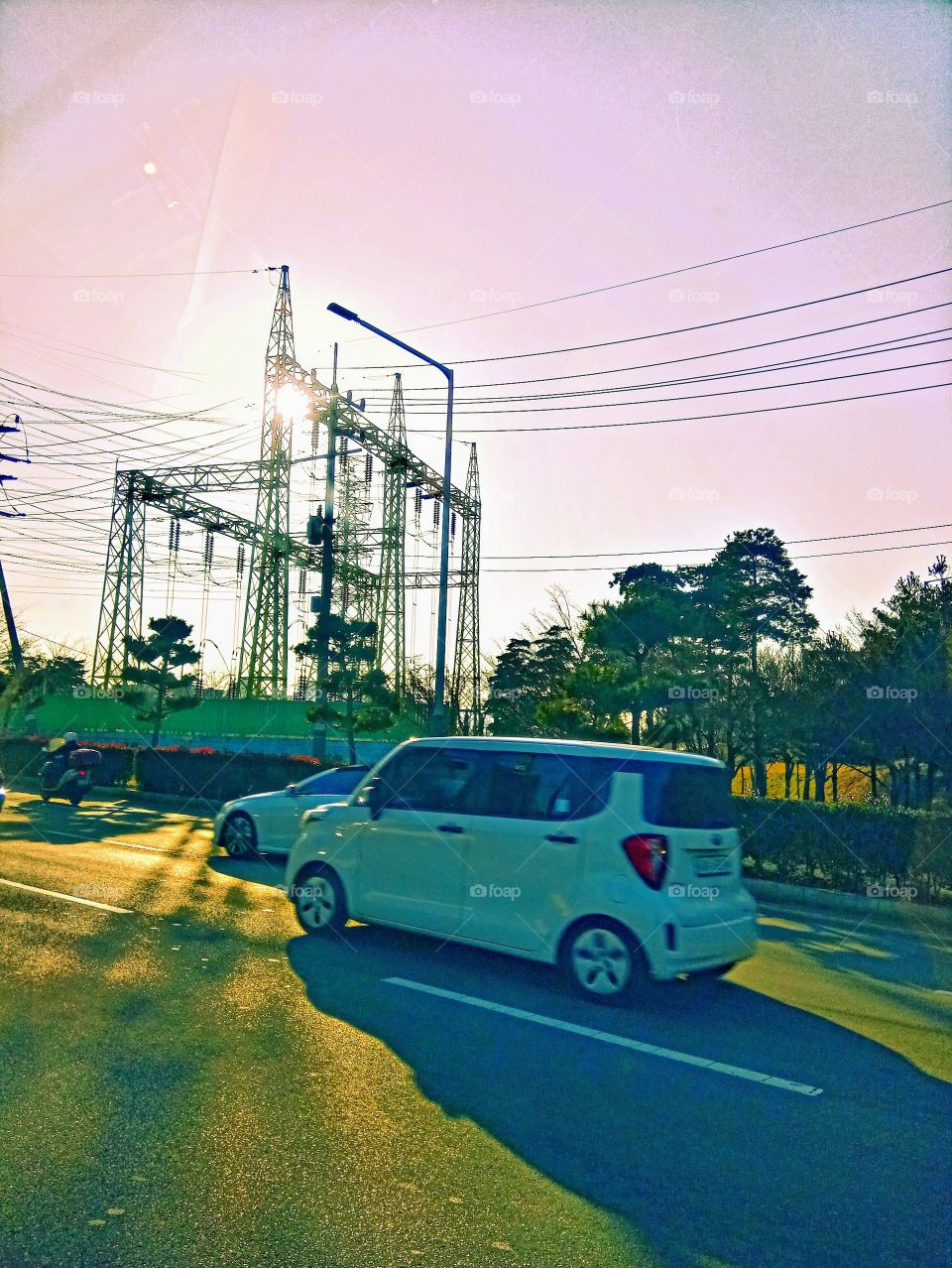 The tall towers with lots of electricity wires that's connected to it and the Beautiful sunlight in the clear sky behind it.
The Cars passing by on the wide Road and the lots of trees beside the road.