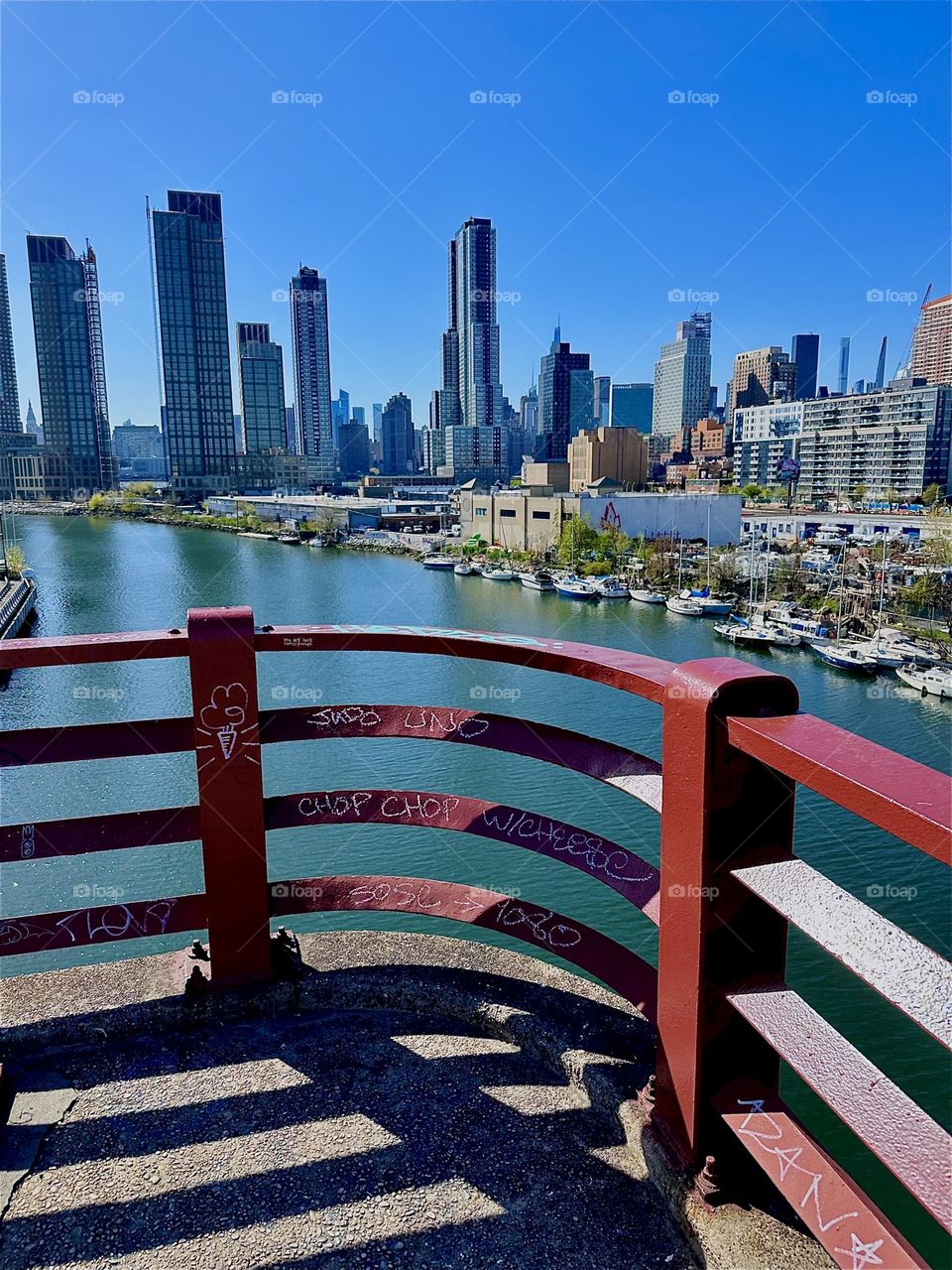 This is “Newtown Creek” with its many boats seen through the red metal railing of the “Pulaski Bridge” that connects “Greenpoint”, Brooklyn to LIC, Queens. In the distance across the “East River” we see “Manhattan”. 2024. Hypnotic Productions