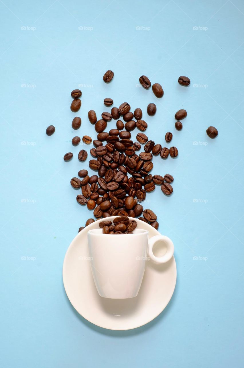 Directly above view of spilled coffee beans and white coffee cup isolated on blue background.