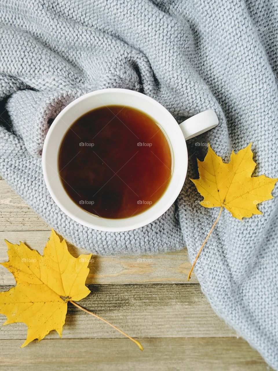 A flatlay style photo of a cup of tea wrapped in a sweater and yellow leaves on a wooden background