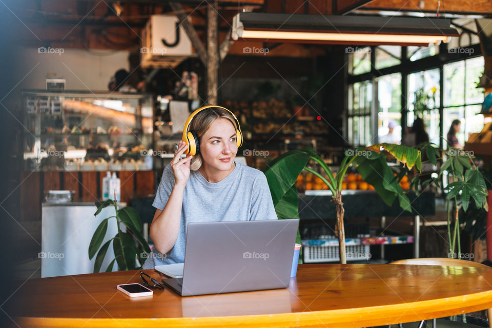 Young blonde woman in yellow headphones and grey t-shirt working on laptop in cafe