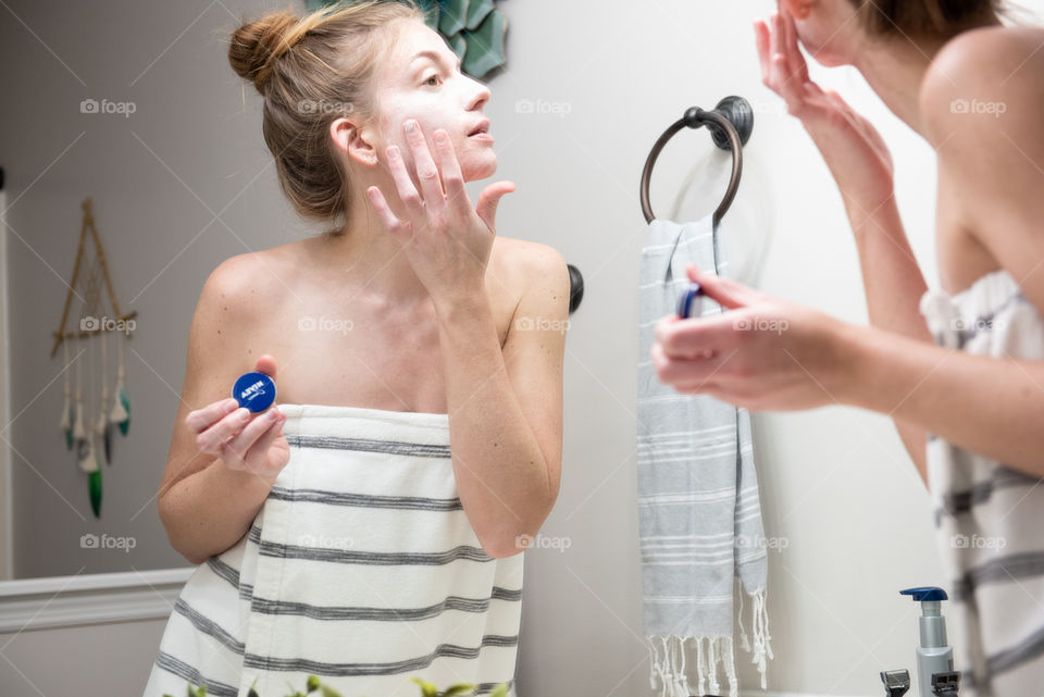 Young millennial woman applying Nivea cream to her face in front of a bathroom mirror
