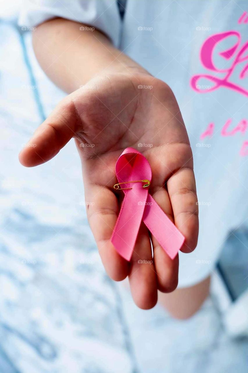 Photo showing part of the silhouette of a person wearing a white tunic with pink markings and an arm presenting in the palm of its hand a breast cancer awareness pink satin ribbon held by a gold nanny pin, over a blurred marbled white floor.