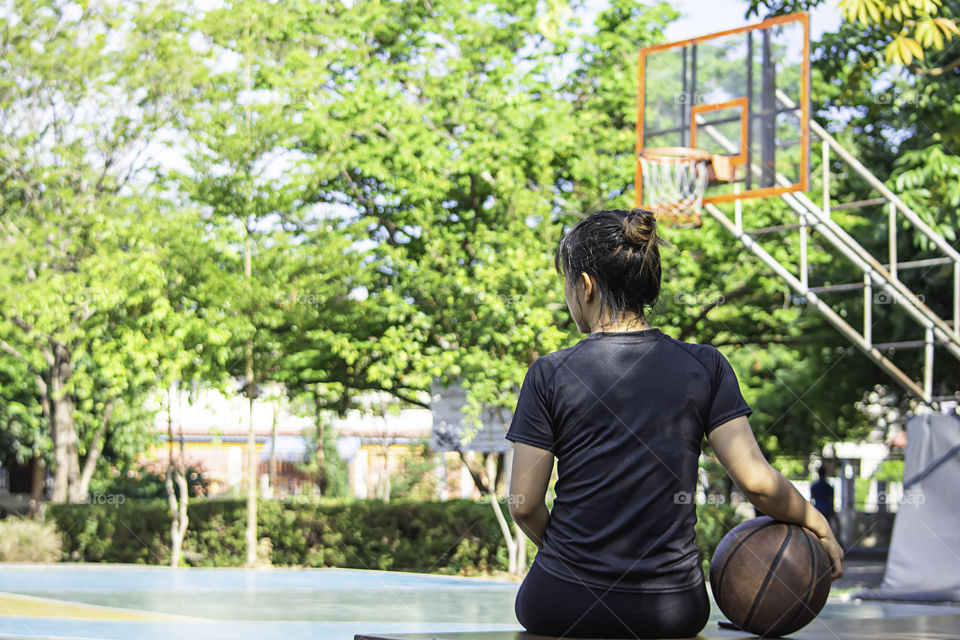 Asian Woman holding basketball sitting on a wooden chair Background blur basketball hoop and tree in park.