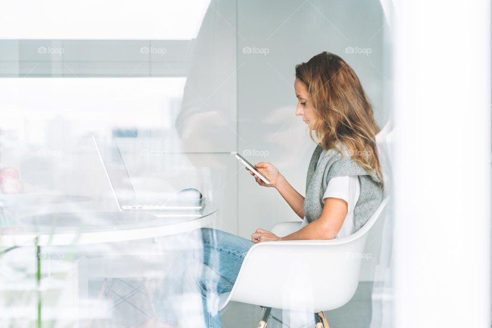 Young adult forty years beautiful blonde woman with long hair working on laptop using smartphone sitting on kitchen at home, remote work