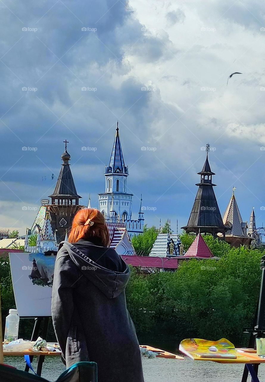 Triangles.  On the shore, a woman draws a landscape.  Among the trees are triangular domes of towers, over which dark clouds have gathered and a bird flies