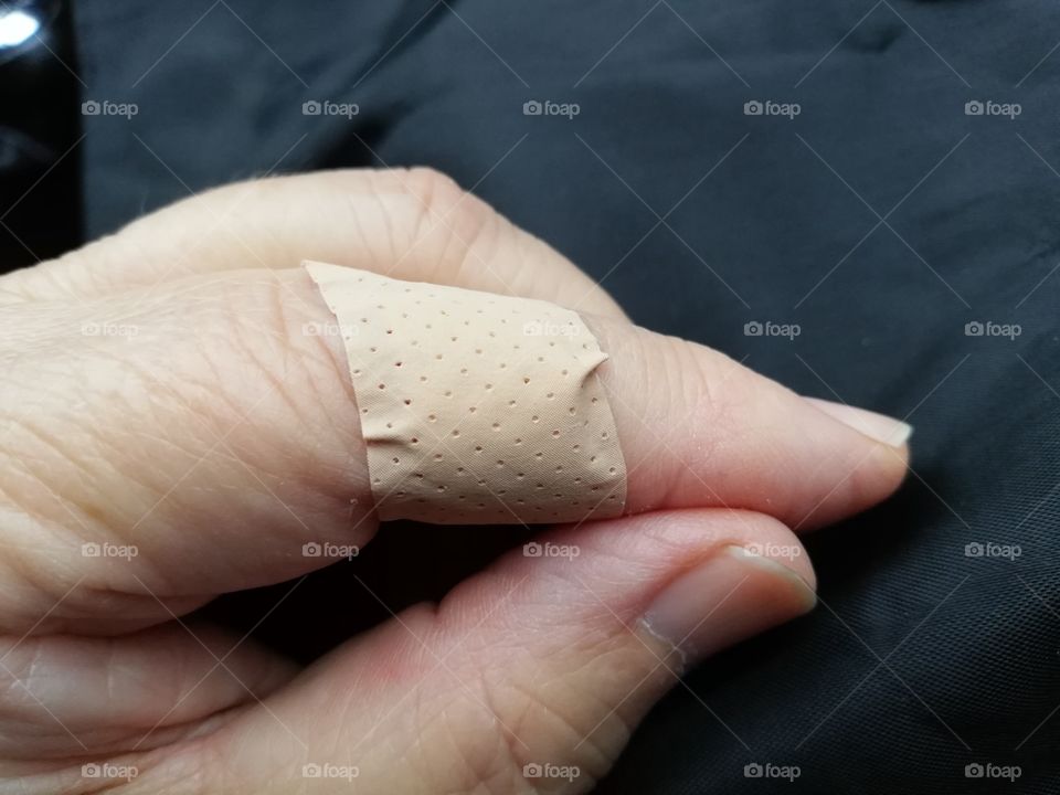 The index finger of the left hand between joints is wounded. The beige plaster pierced with small holes is set around the finger. The fair skin is wrinkled, the cuticle on the thumb broken, hand resting on a black fabric.
