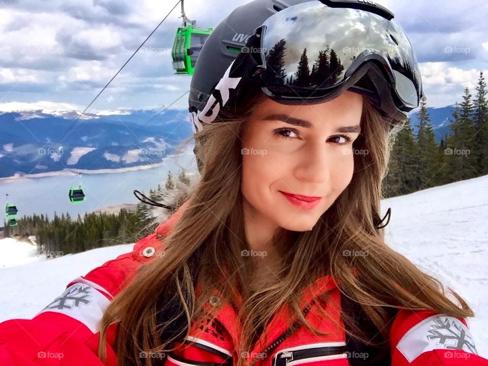 Selfie of pretty young woman wearing Uvex ski glasses and Uvex ski helmet on the slope with ski lift in the background