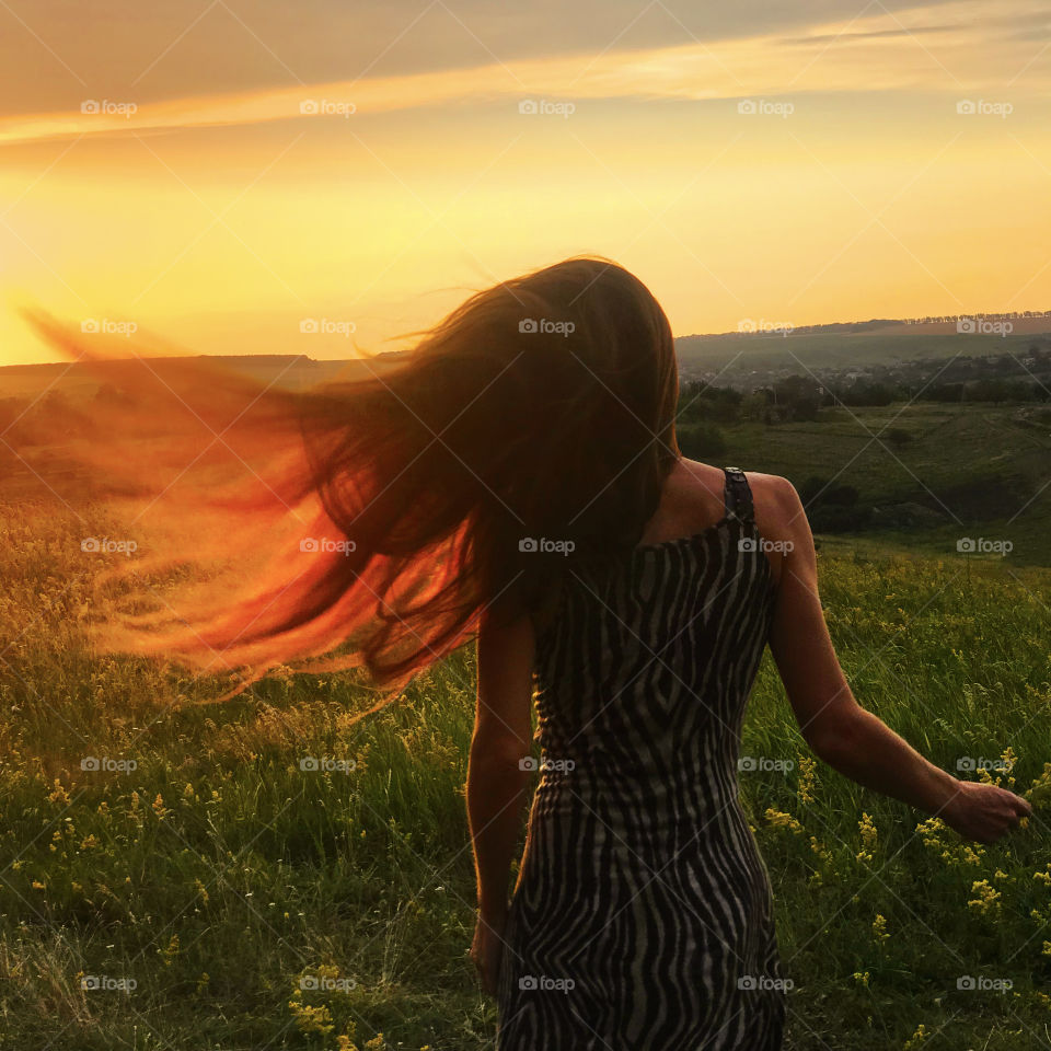 Young woman catching orange evening sunlight by her long hair in the summer field 