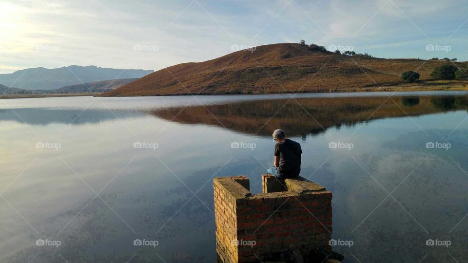 Solitude... lone boy sitting watching a calm lake with mountains in the backround at dawn