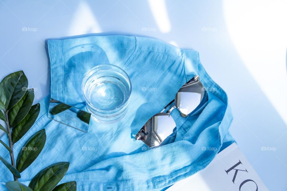 Items and image are selected and prepared for travel. A blue shirt, a glass of clean water and sunglasses, reading magazines on a blue background.