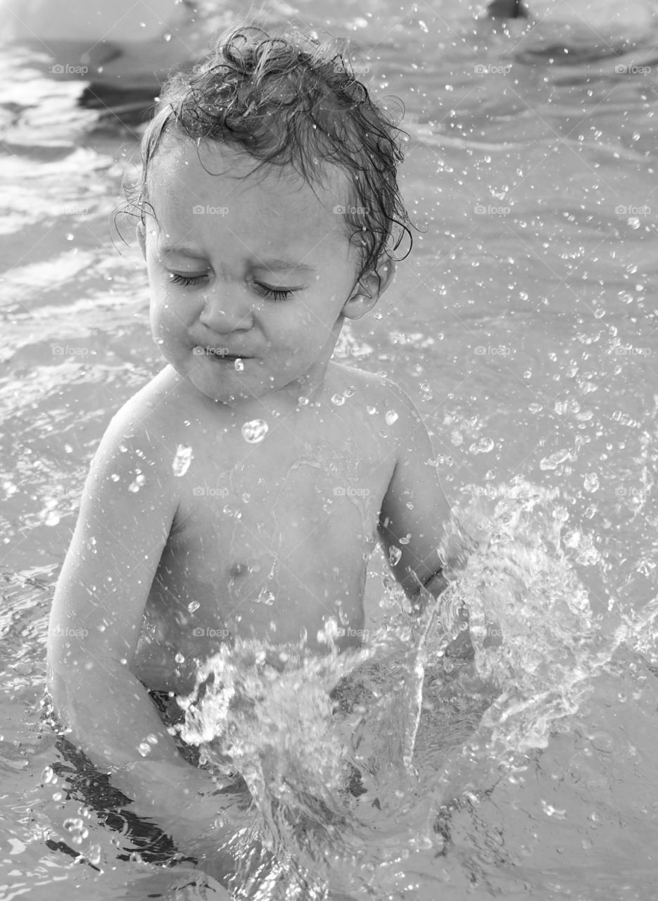 A little toddler boy closes his eyes and turns his head after splashing water in a swimming pool. 