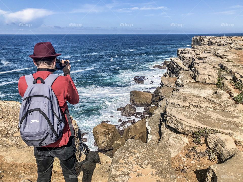 A tourist with a backpack takes photos of the rocky coastline at Peniche in Portugal 