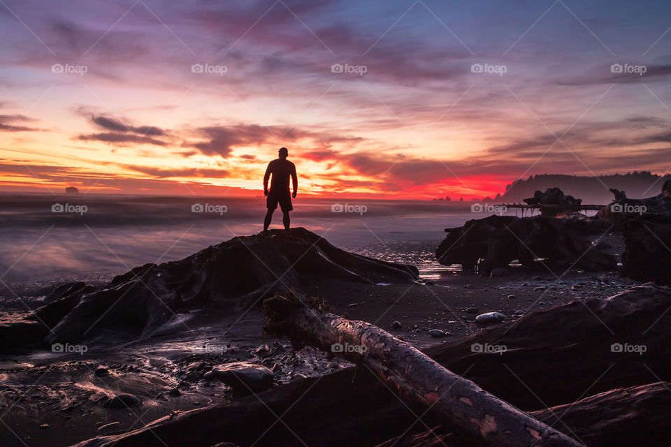 Silhouette of a person standing on driftwood, watching as the tide comes in on a rugged coastline under a beautiful vibrant sunset.