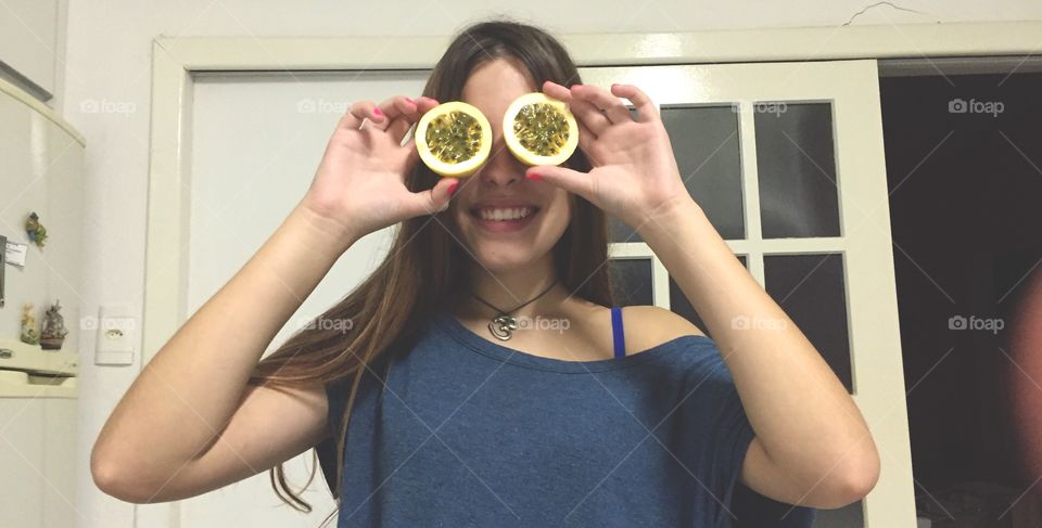 Happy woman holding slice of passion fruit on her eyes