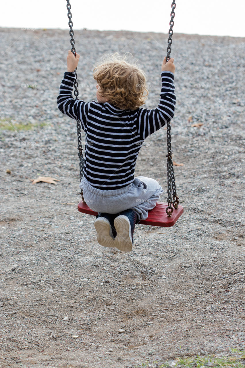 a boy on a red swing seen from the back looking towards the sea