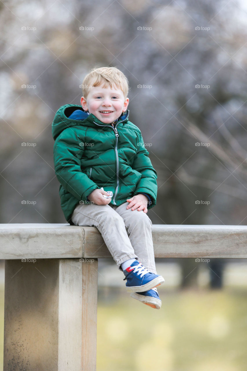 Little boy with blonde hair smiling and sitting on park bench in green coat 
