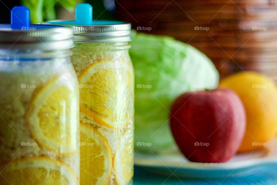 Fermented cabbage, apples and oranges in quart-size mason jars with blue silicone fermenting lids, a cabbage, an apple, and an orange on a white plate on a blue placemat in the background