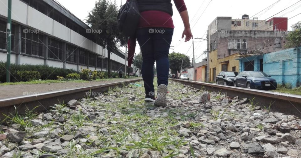 mujer caminando en las vías del tren dentro de la ciudad, con piedras y hierbas, vistiendo pantalón de mezclilla y tenis
