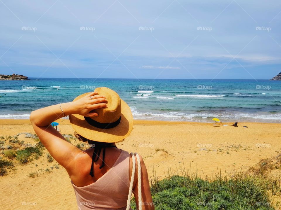 woman from behind with straw hat looks at the beach and the sea from a cliff