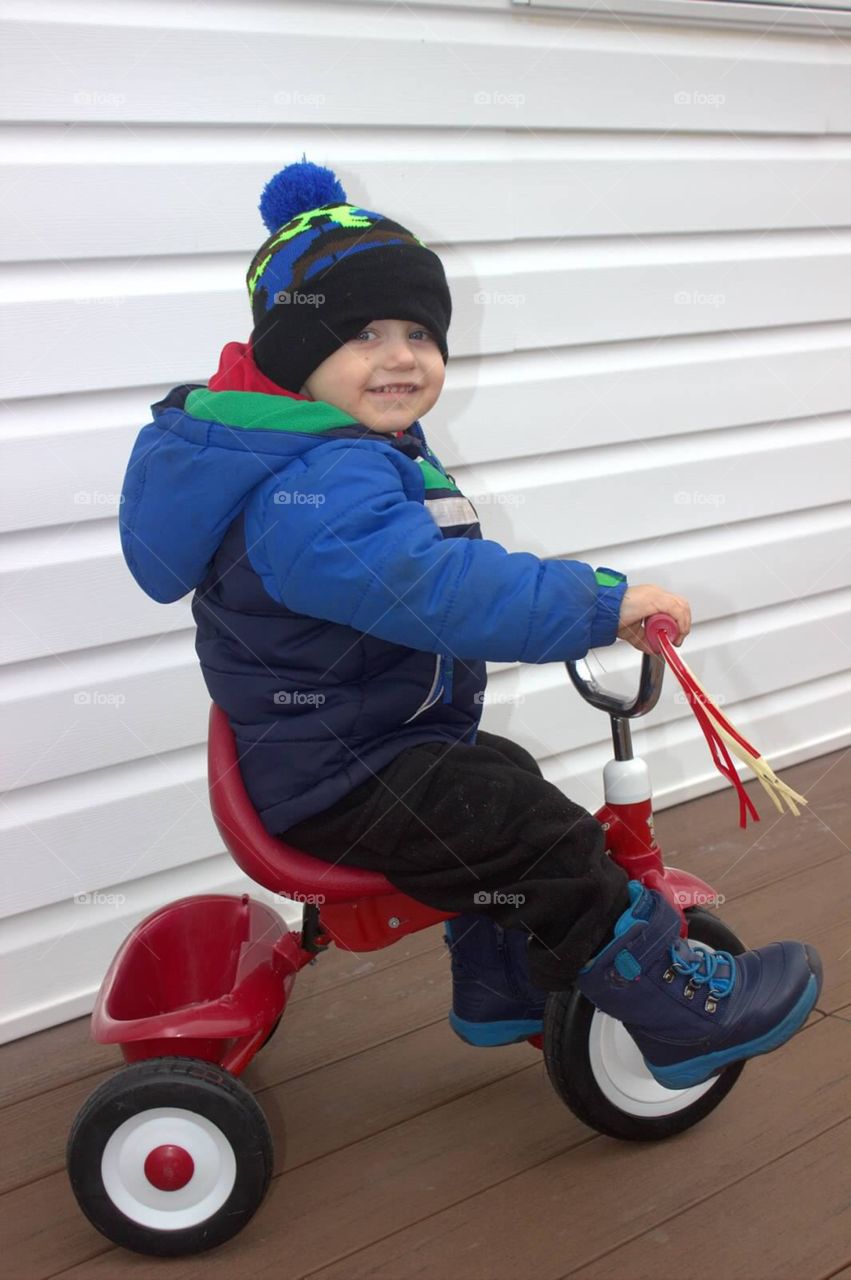 A two-year-old little boy riding his tricycle on a cold winter day. 