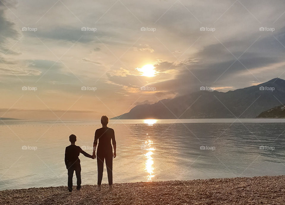 Silhouettes against the setting sun.  Mom with a baby boy are standing holding hands on the seashore and watching the sunset.  Summer family vacation at sea