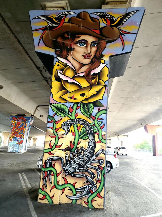 Colorful street art on highway bridge underpass cement pillar. A cowgirl blooming out of a yellow rose with a scorpion on the ground beneath and birds on each side of her hat.