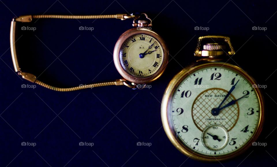 Flat lay of a woman’s gold vintage wristwatch with a slim expandable gold band and a man’s gold vintage pocket watch on a black surface