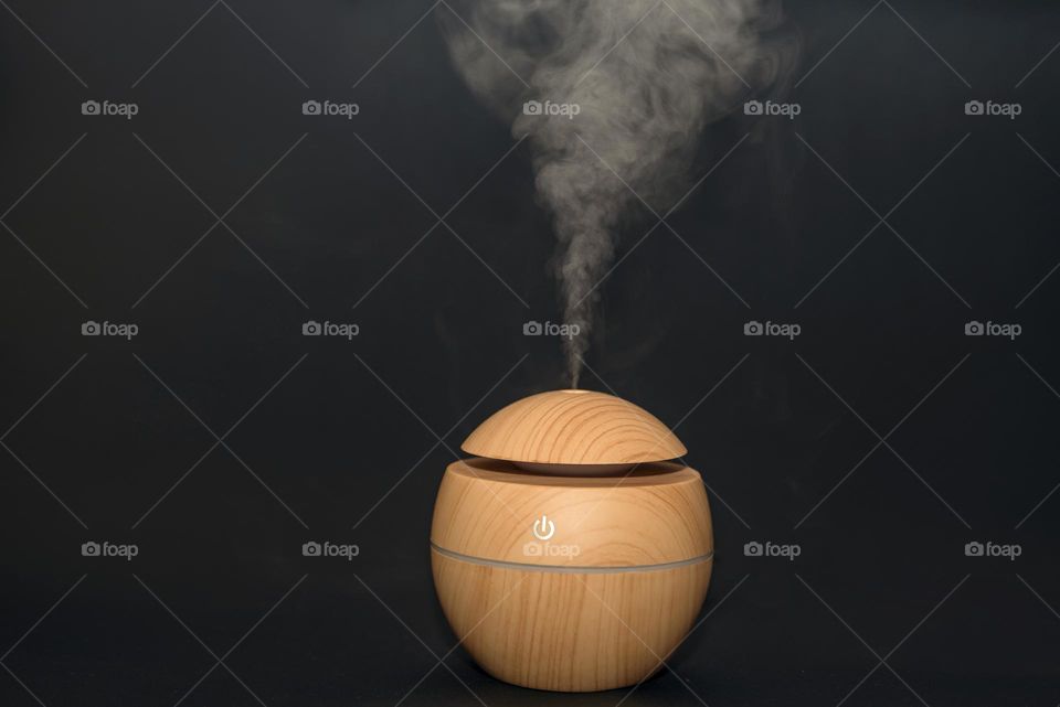 An electric ultrasonic humidifier emits atomized water droplets against a dark background. The device is round in shape with a wooden body.