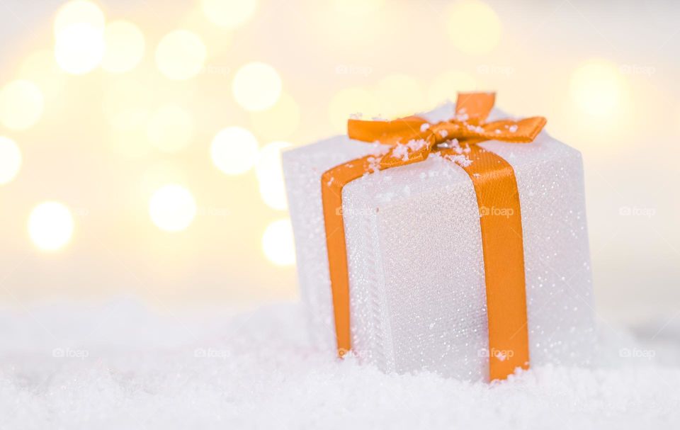 One Small white gift box with orange ribbon stands on the right in the snow with small copy space on the left on a yellow background with bokeh and lights, side view close-up. Holiday gifts concept.