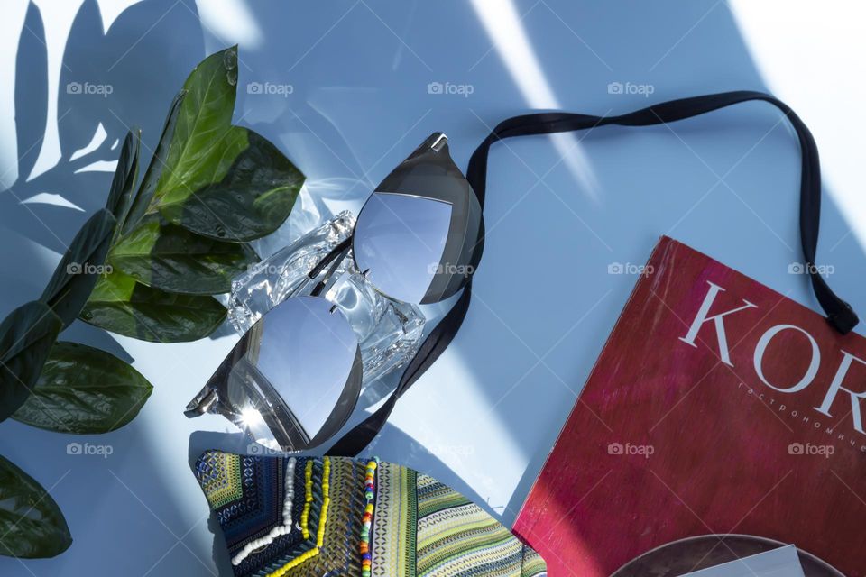 Items and image are selected and prepared for travel. A blue shirt, a glass of clean water and sunglasses, reading magazines on a blue background.