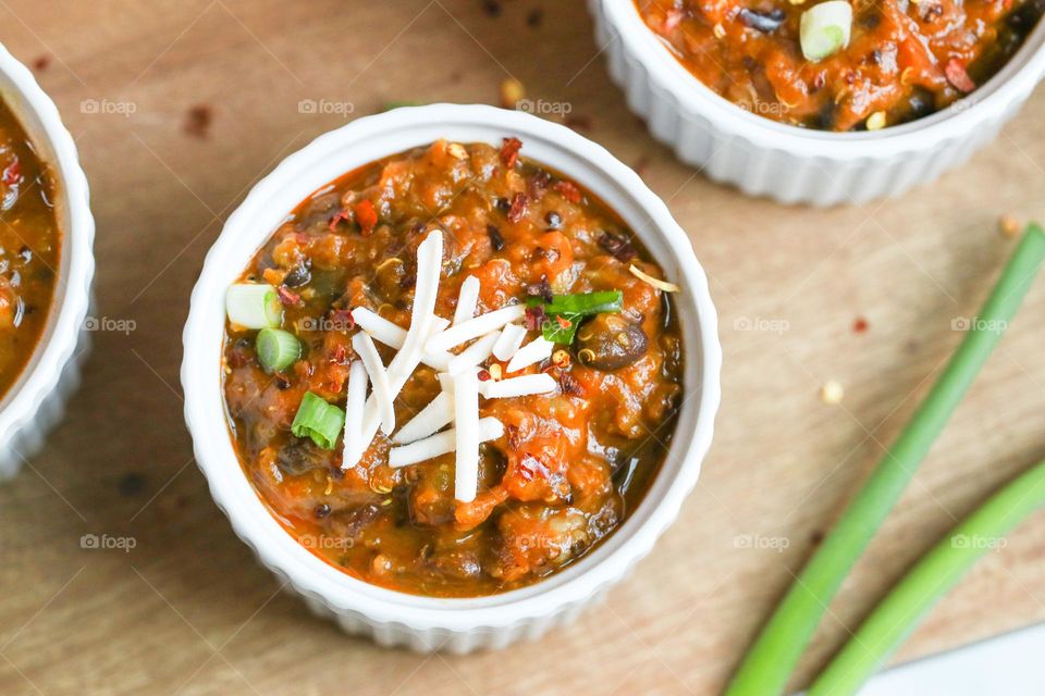 Black bean and sweet potato chili in a white Ramekin. Topped with vegan cheese, scallions, and red pepper flakes. On a wood board.  
