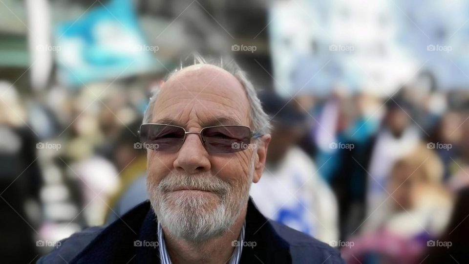 "A father figure to many." A man smiling, looking forward, a blurred crowd behind him. He is unique and special. He is a grandfather, a father, a son, a brother, a partner, a friend, a caregiver, and a good listener to anyone with good will.