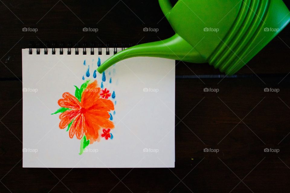 Flat lay watercolor crayon drawing in a mixed media notebook of a large orange flower being “watered” by a green watering can so that on the watered side the colors are blending, dark wood background