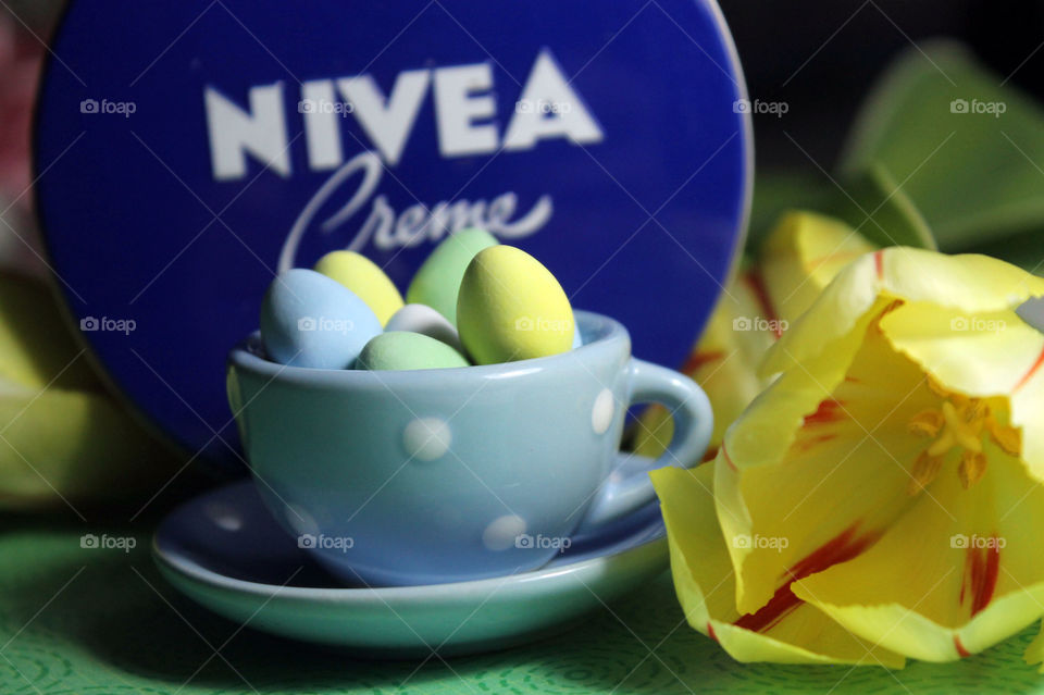 A closeup of a blue container of NIVEA Creme. The scene also has a small ceramic blue polkadot teacup & saucer filled with pastel candy coated chocolate Easter eggs & yellow tulips all on a green  base. 