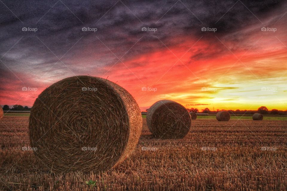 Bails of hay sit in a farmers field at sunset with a dramatic sky overhead.