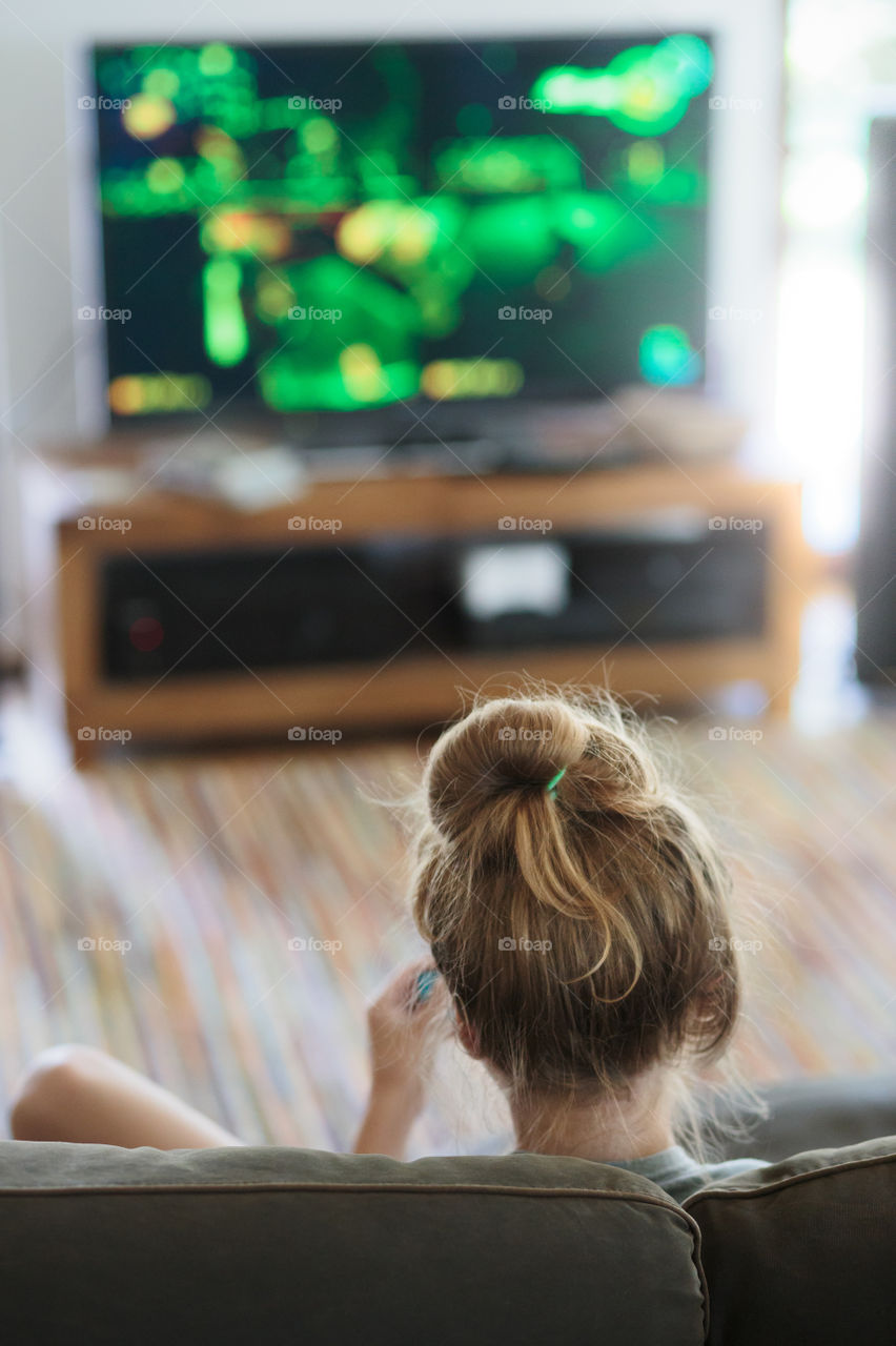 Back view of young girl playing video game sitting on sofa at home