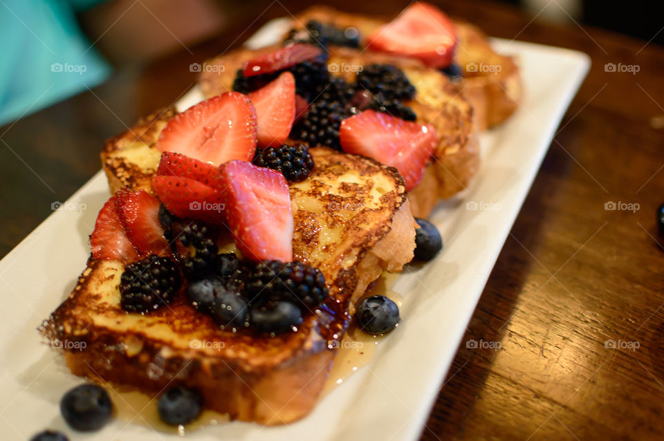 Beautiful sliced French toast on platter piled with a mountain of strawberry, blackberry, blueberry fruits and maple syrup healthy brunch or breakfast photography 