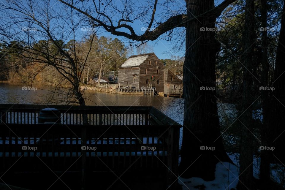 Late afternoon view with the sunshine falling upon the old gristmill with snow on the roof across the millpond, with the silhouettes of a barren oak tree and an observation deck at Yates Mill County Park in Raleigh North Carolina.