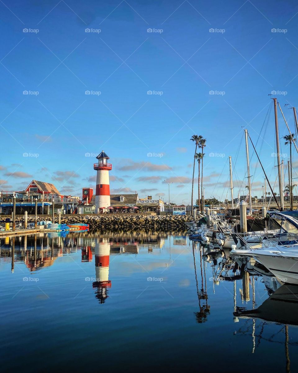 A striped lighthouse is reflected in the still waters of the marina full of sailboats