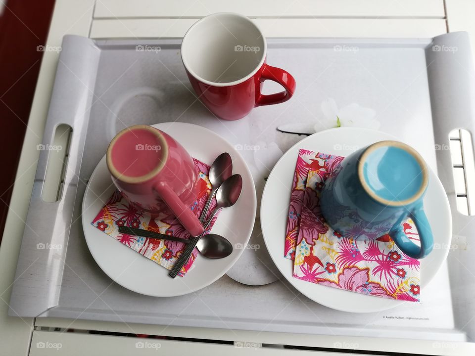 Three mugs on a plastic tray. Two of them upside down on the patterned napkins on the white plates. On another plate three metallic ornated teaspoons.