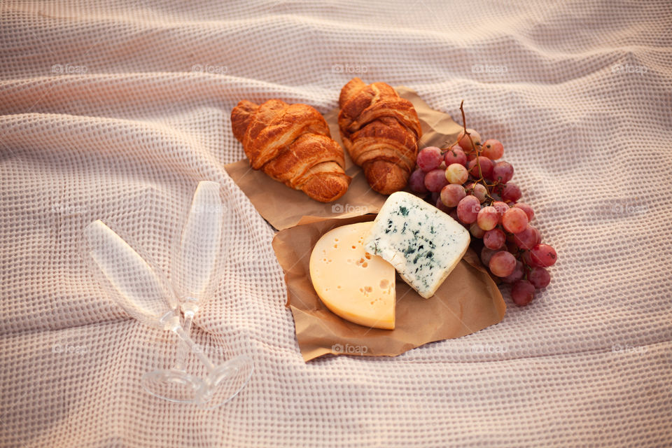 Romantic picnic for two at sunset. Two pieces of cheese, grapes, two croissants and two empty glasses are on the blanket. Close-up photo