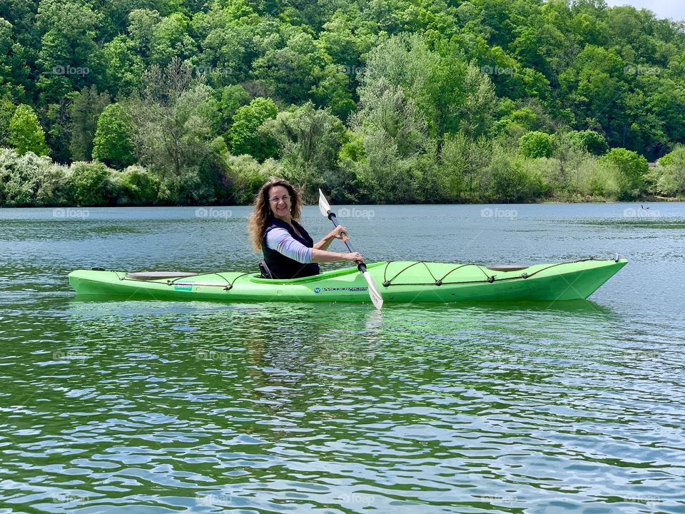 Woman in green kayak with paddle on lake with backdrop of trees.