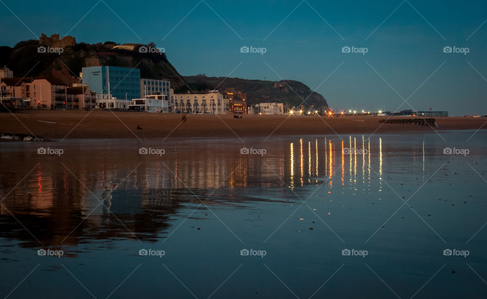 A view of Hastings seafront and cliffs from the beach, the tide is out and the lights reflect on the wet sand.