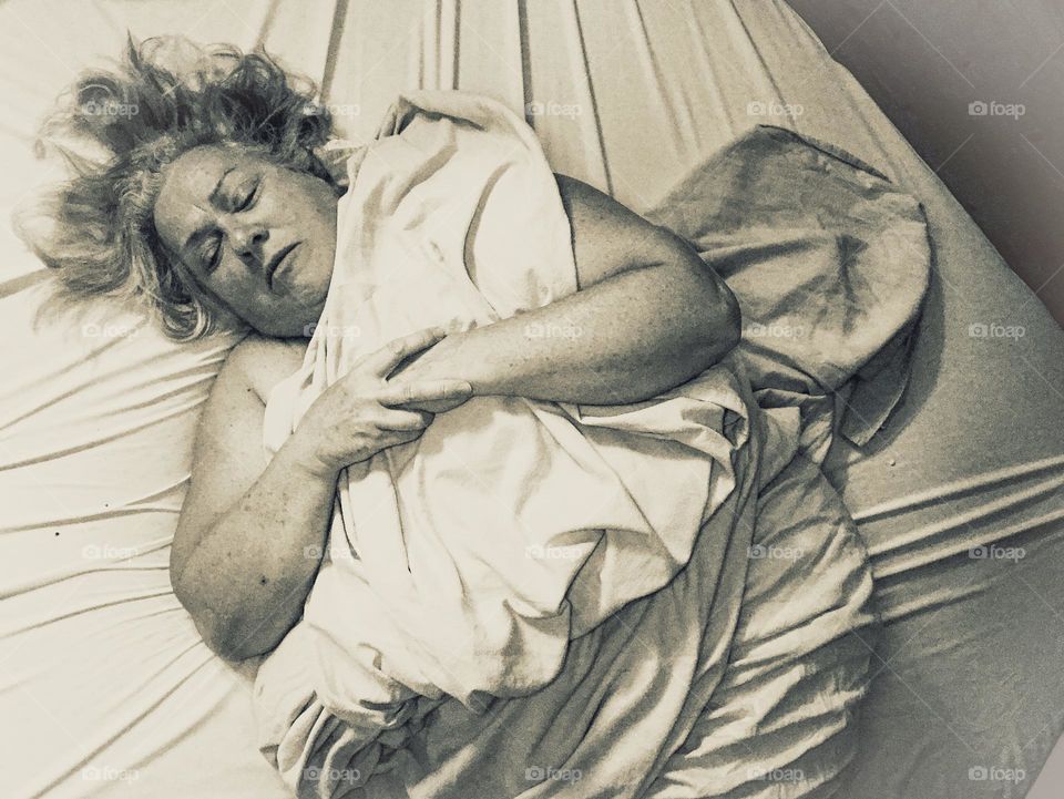 Silver tone self-portrait of a woman lying on a bed tightly bundled in a sheet.  Her hair is messily splayed around her face, her eyes are closed and her hands are tightly clenched together. Her whole body appears tense and rigid. 