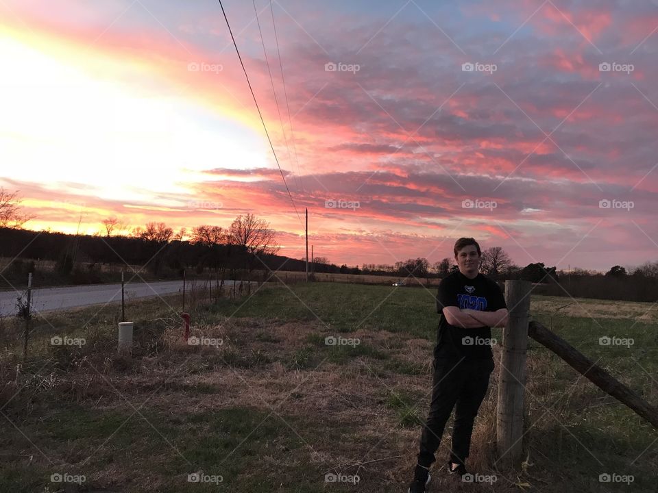 Young man standing by fence post with beautiful sunset in the background.