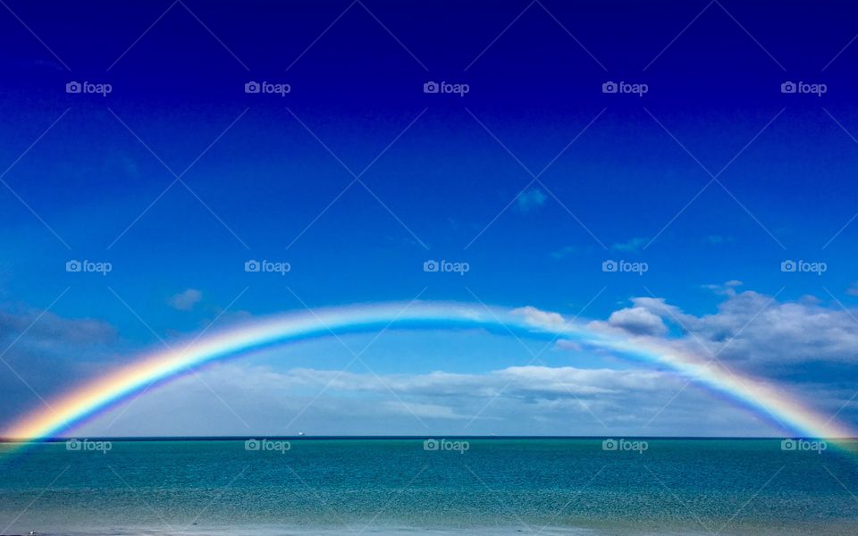 Full colourful rainbow with faint double arching and stretching over the ocean horizon and through the aquamarine turquoise blue water, on brilliantly sunny blue sky and light cloudy day, South Australia, Spencer Gulf Eyre Peninsula