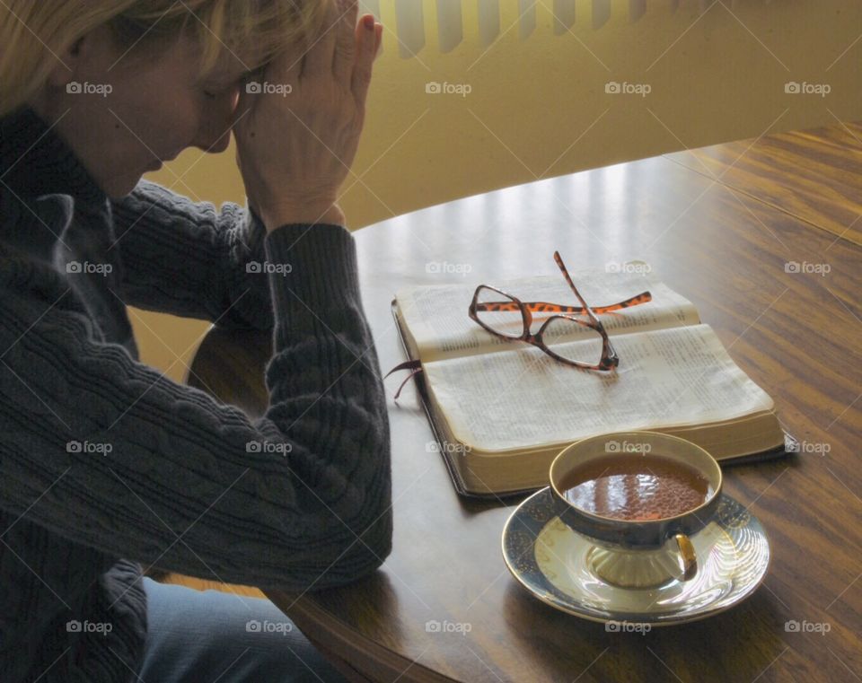 Woman with head bowed in prayer, Bible open with eye glasses sitting on it and a cup of tea beside the Bible